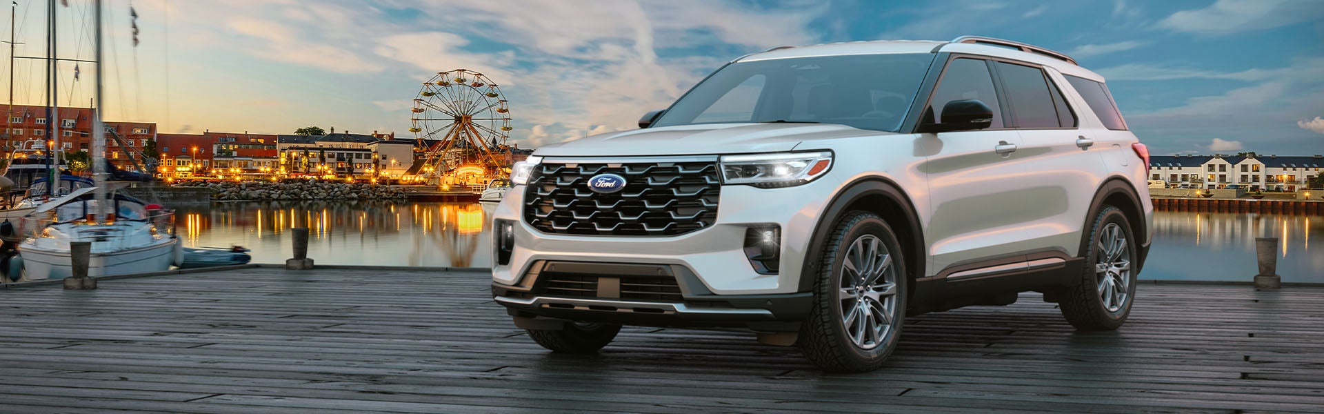 White Ford Explorer SUV parked on a wooden pier with a town, Ferris wheel, and boats across the water at sunset.