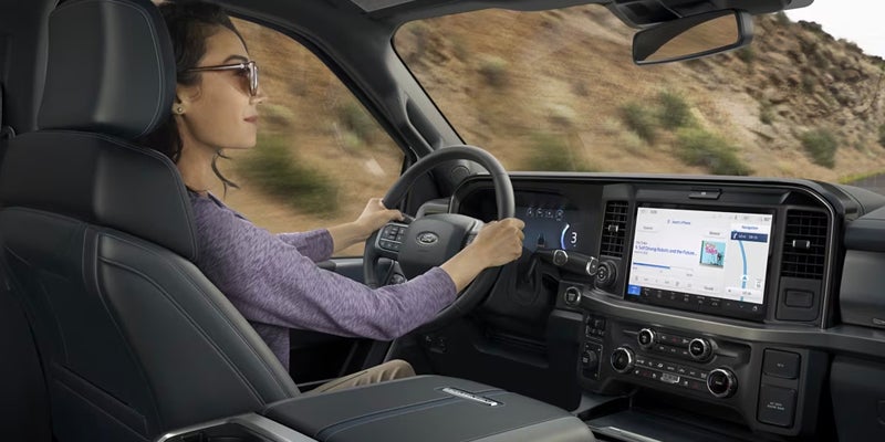 A woman with sunglasses drives a modern truck, its dashboard displaying a navigation map and music playing.