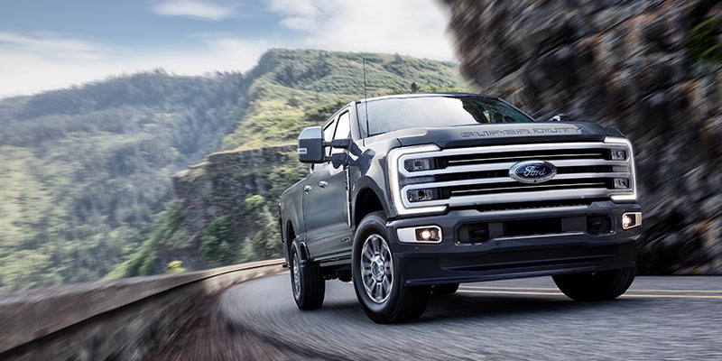 A dark gray Ford Super Duty pickup truck driving along a winding mountain road with green, forested hills.