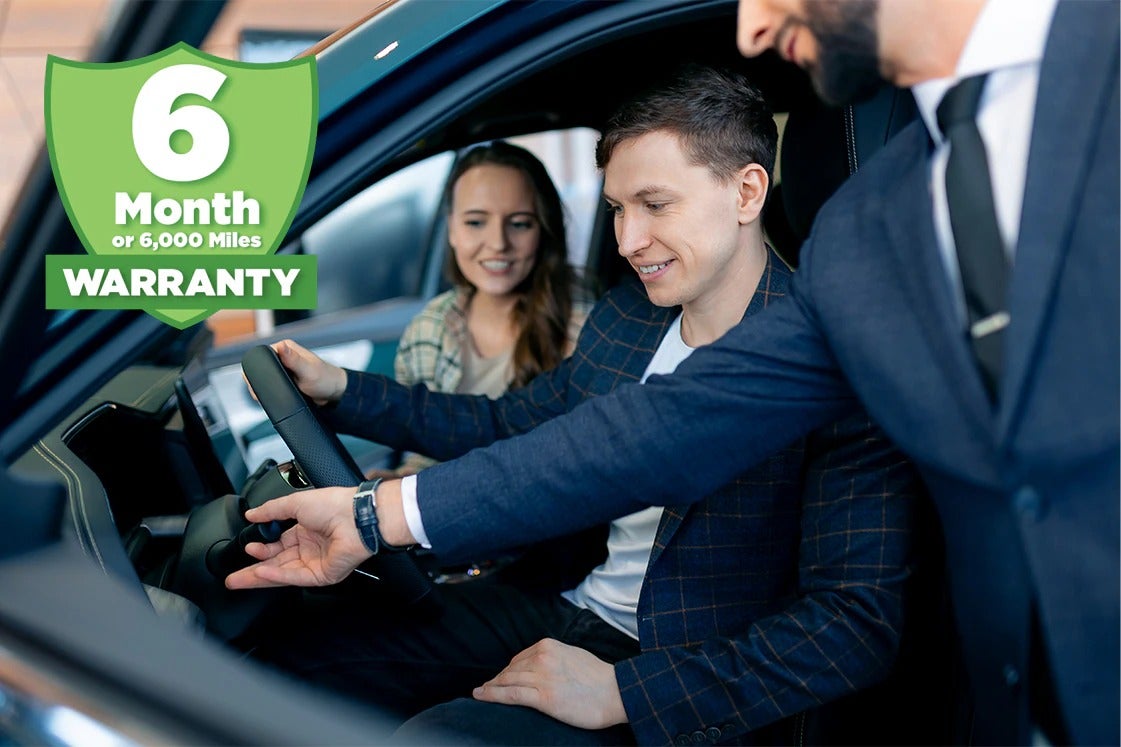 Man and woman in a car with a salesman, a warranty badge for 6 Month or 6,000 Miles Warranty overlayed.