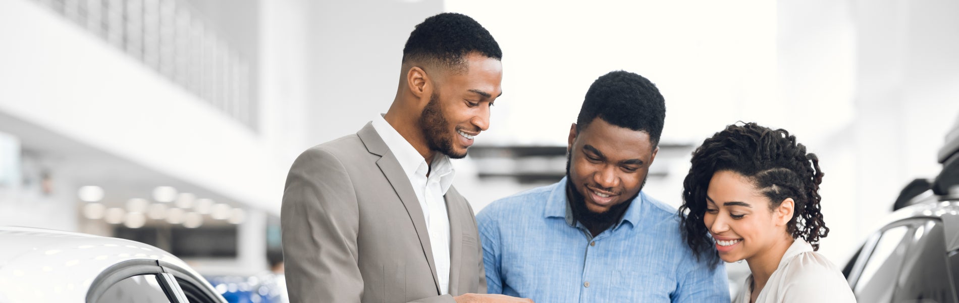 A smiling car salesman helping a young couple at a dealership, showing them information on a tablet.