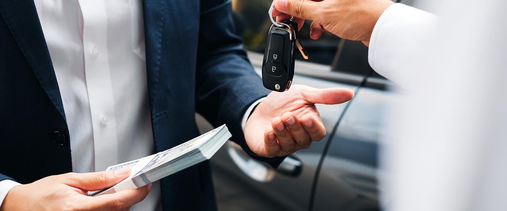 A person in a suit holds a stack of money while another person hands them car keys, with a car in the background.