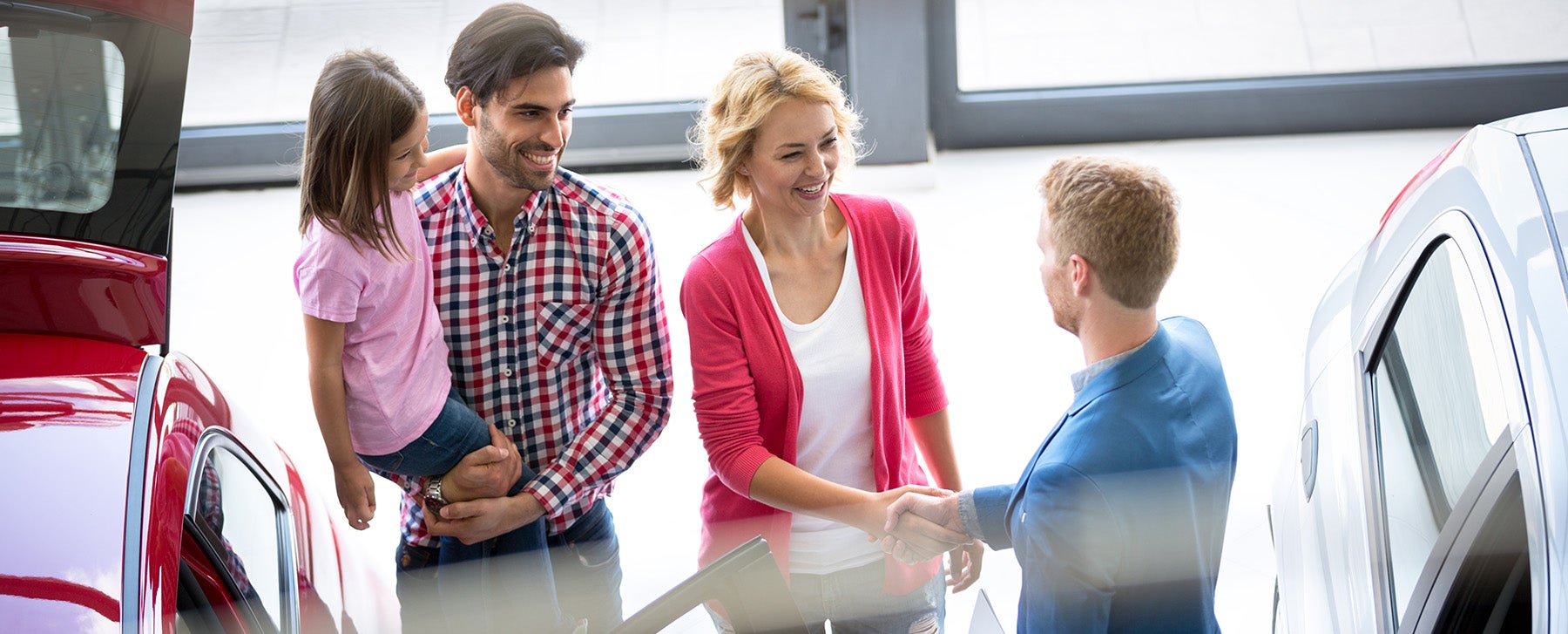 A family with a young girl shaking hands with a car salesperson in a showroom, with new cars around.