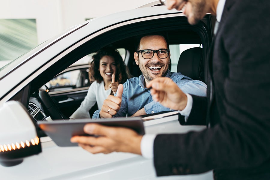 Happy couple in a car giving thumbs up to a smiling salesman