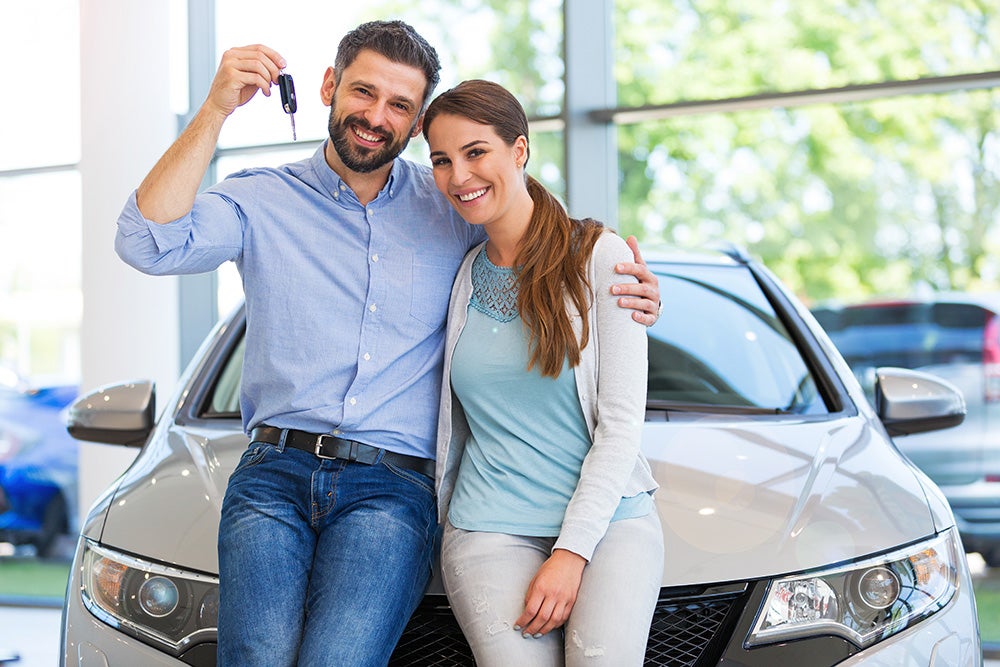 Smiling couple sitting on car hood holding keys in a dealership showroom